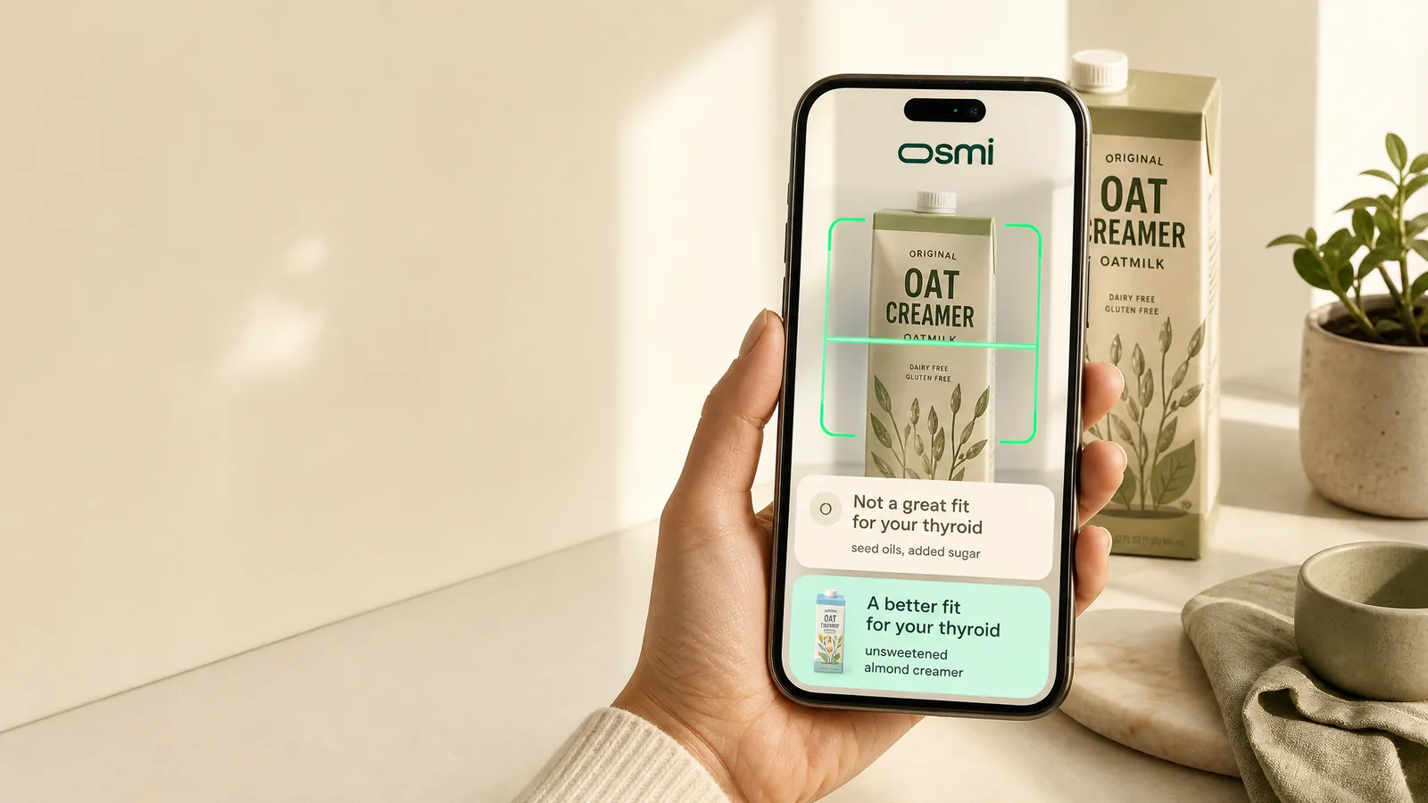 A hand holds a phone scanning an oat creamer in a cream kitchen. On screen, osmi reads: not a great fit for your thyroid, and suggests a better fit, unsweetened almond creamer.