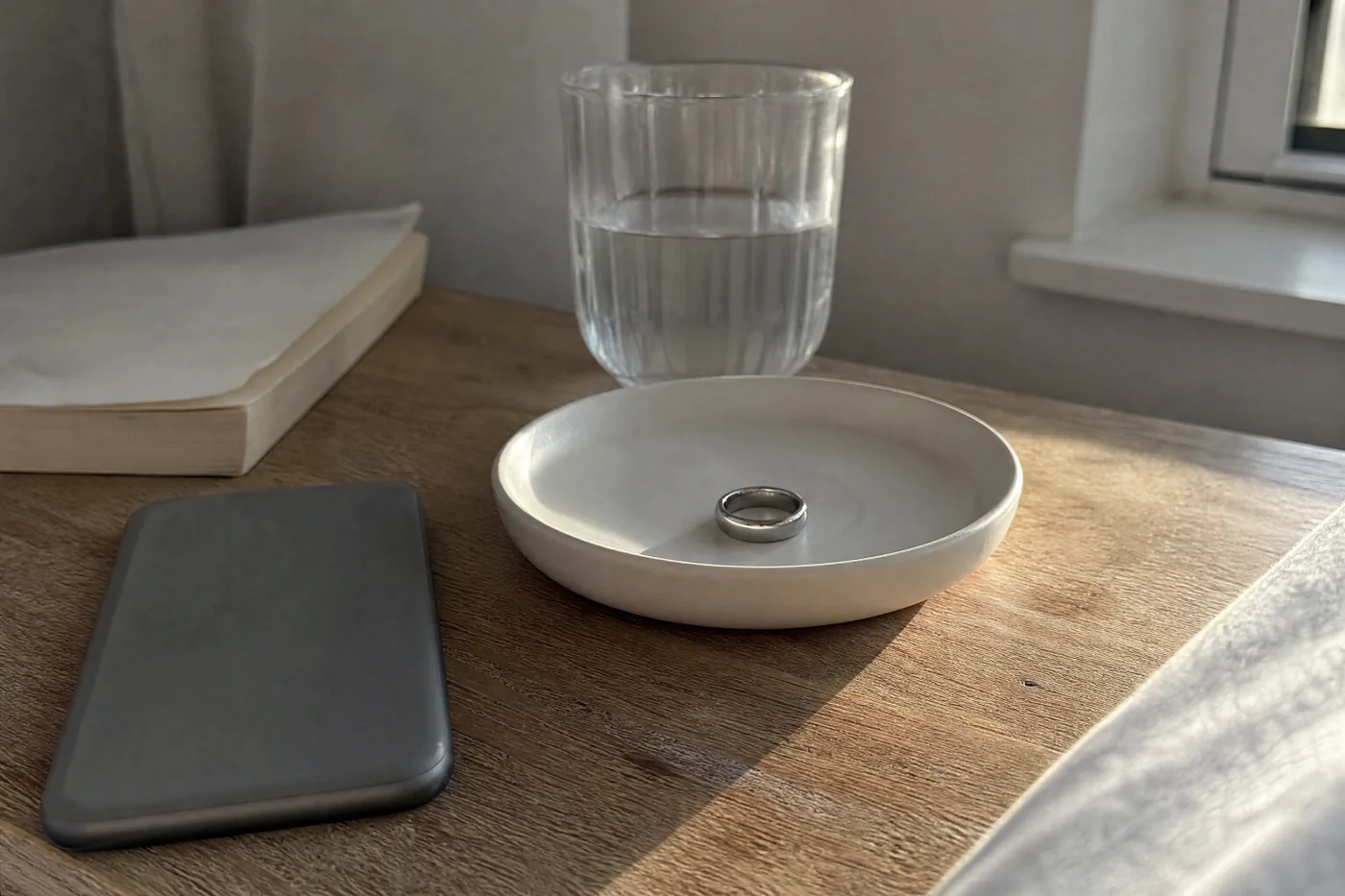 A plain silver ring in a small ceramic dish beside a phone lying face down on a bedside table.