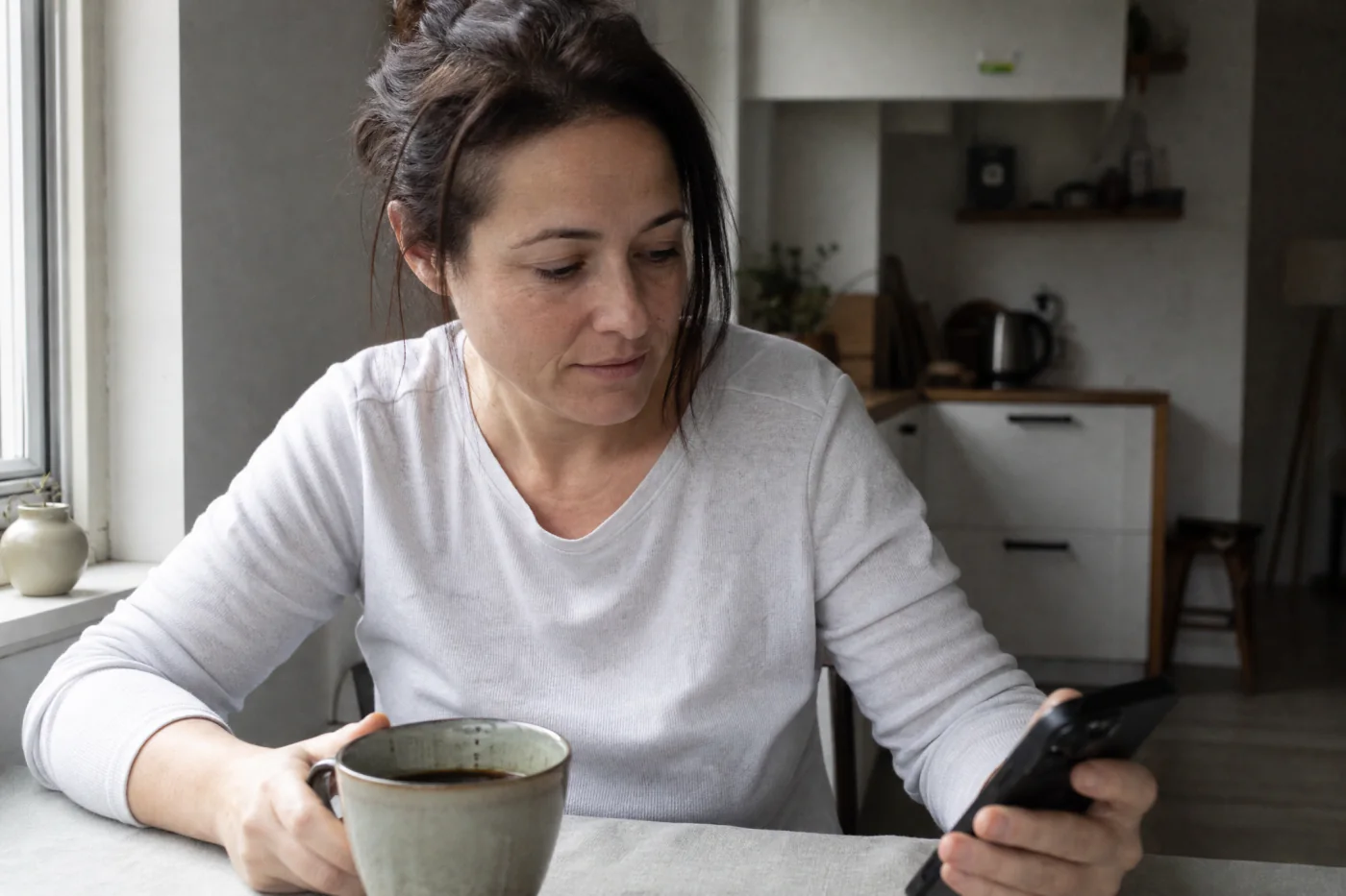 A woman at a kitchen table in the morning, one hand on her coffee and one on her phone.