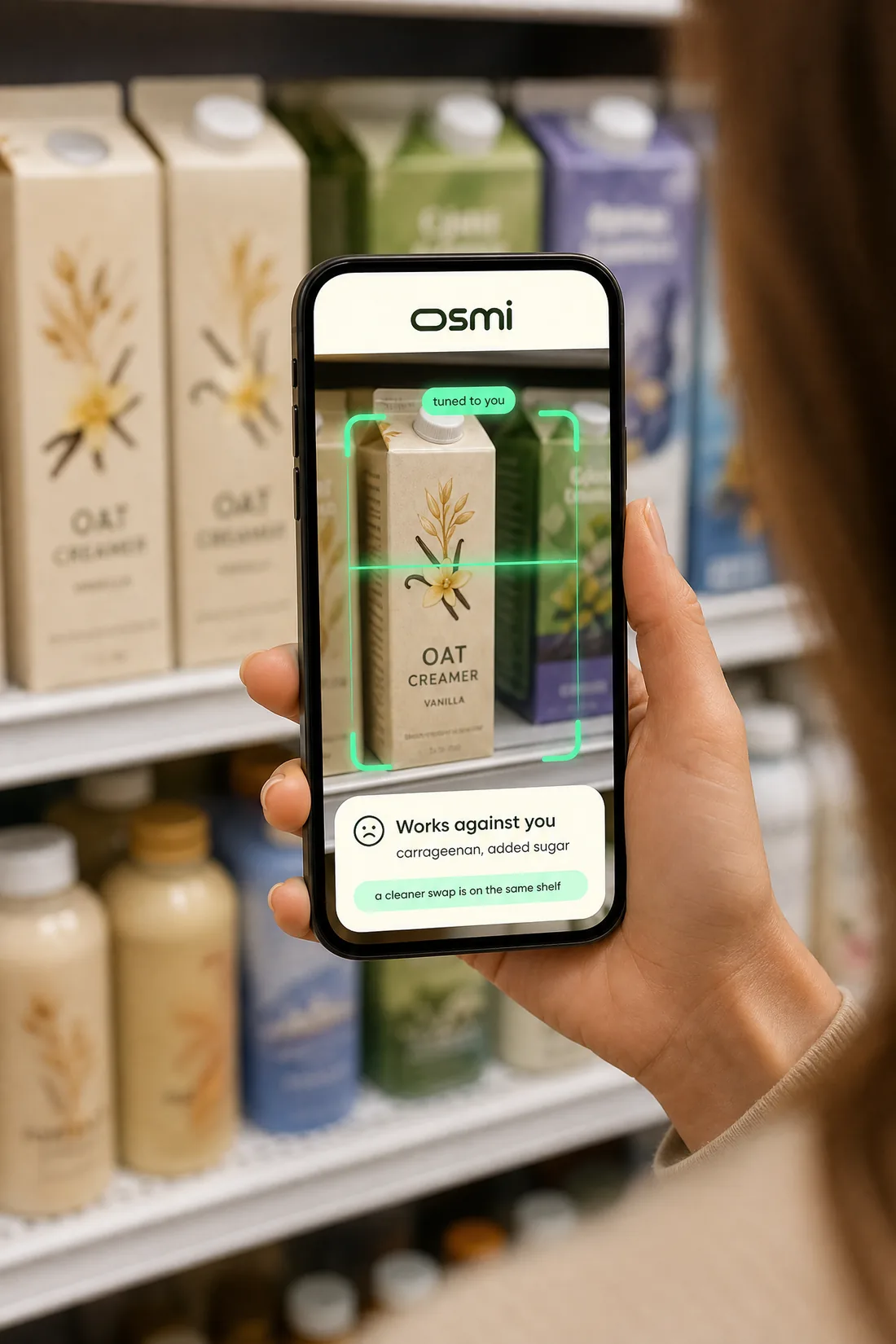 In a grocery aisle, a woman photographs a vanilla oat creamer on the shelf with her phone. On screen, osmi reads it tuned to you: works against you, carrageenan and added sugar, with a cleaner swap on the same shelf.